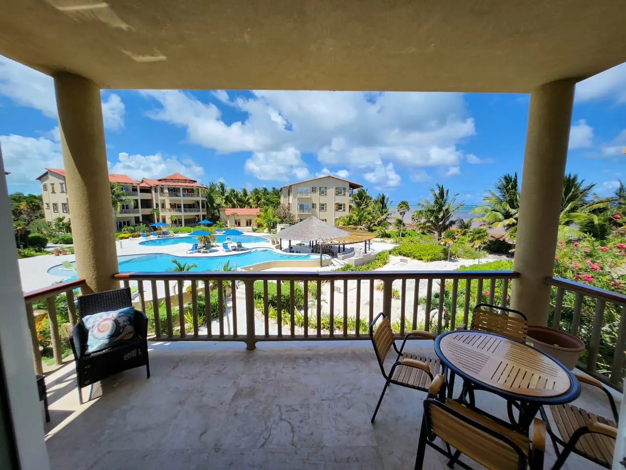 Balcony view of a resort with a pool, palm trees, and buildings under a blue sky with white clouds. Table and chairs on the balcony.