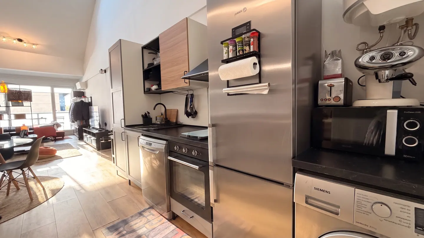 A modern kitchen with stainless steel appliances, dark countertops, and light wood floors. A living area is visible in the background.