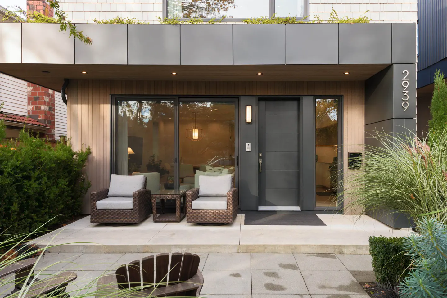 Modern home exterior with a gray door, number 2939, and patio furniture. Bushes and plants surround the entrance.