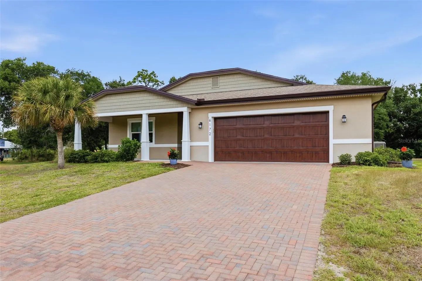Beige single-story house with a brown garage door and a brick driveway. A palm tree is on the left side of the house.