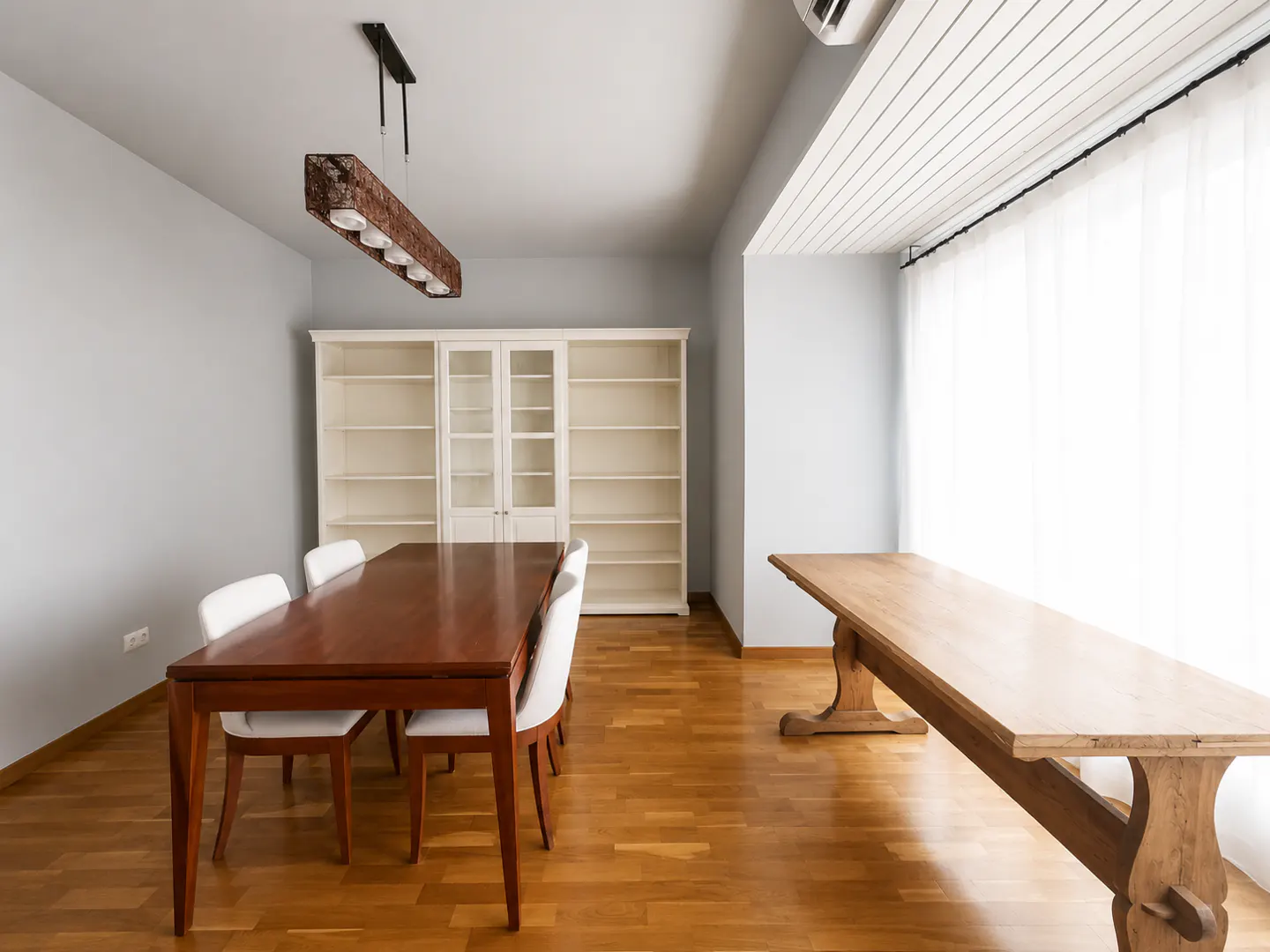 Dining room with a dark wood table, white chairs, a large white bookshelf, and a light wood table on hardwood floors.