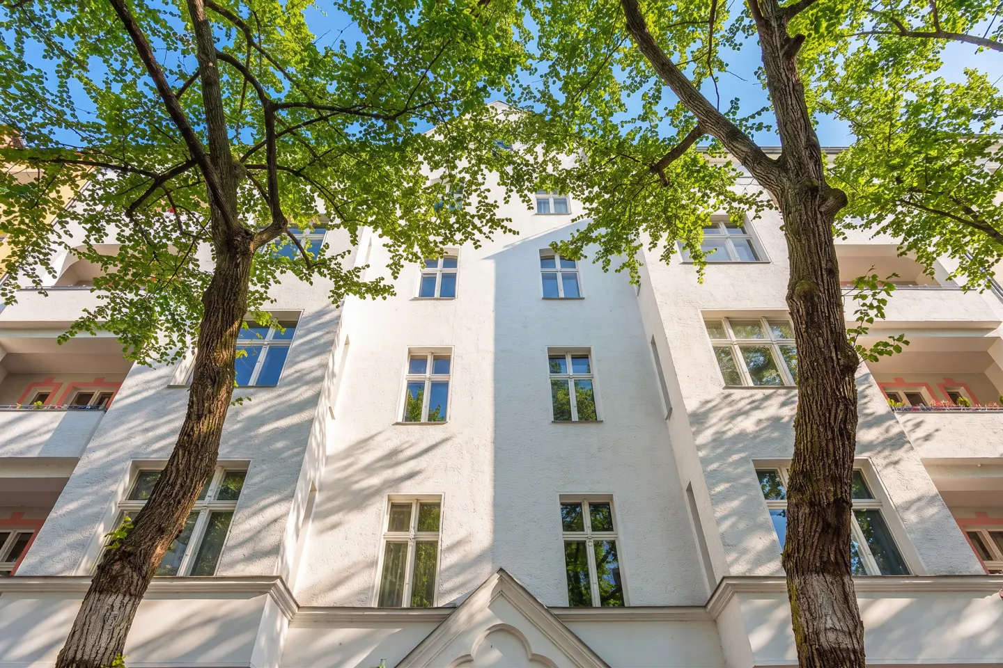 Low angle view of a white apartment building framed by two trees with green leaves against a blue sky.