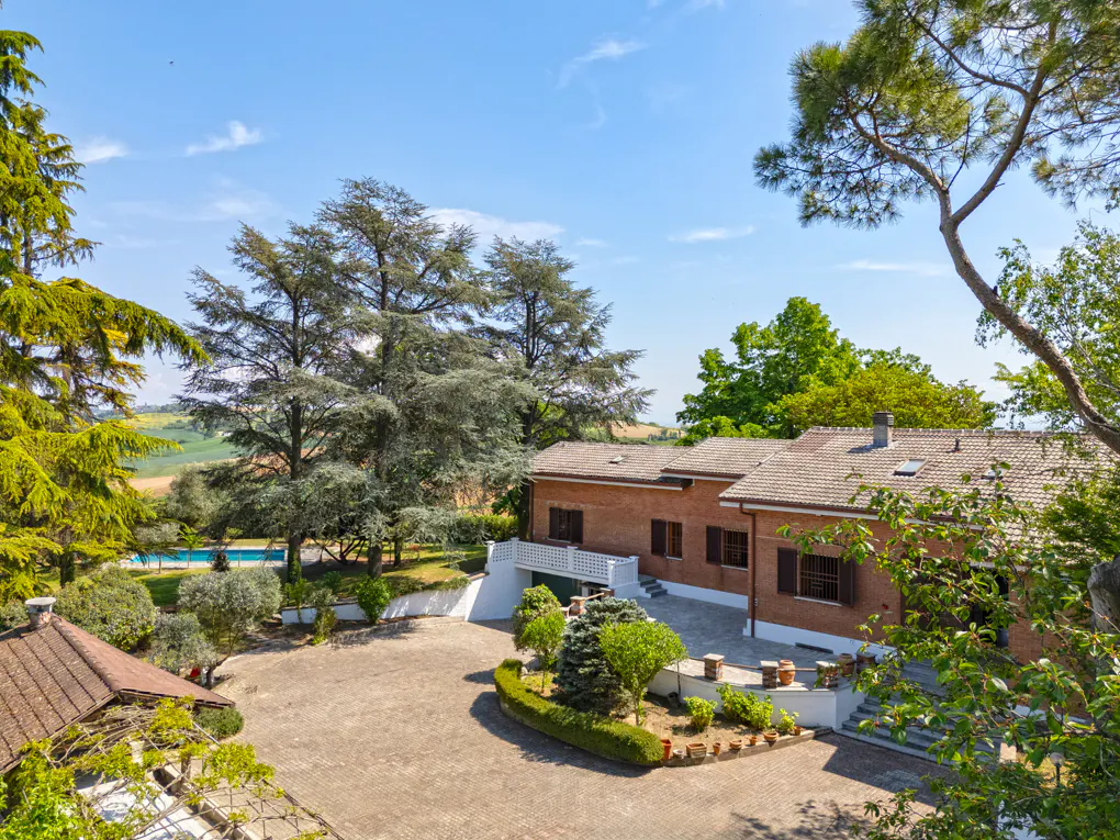 Exterior view of a red brick house with a large paved courtyard, green trees, and a blue pool in the background.