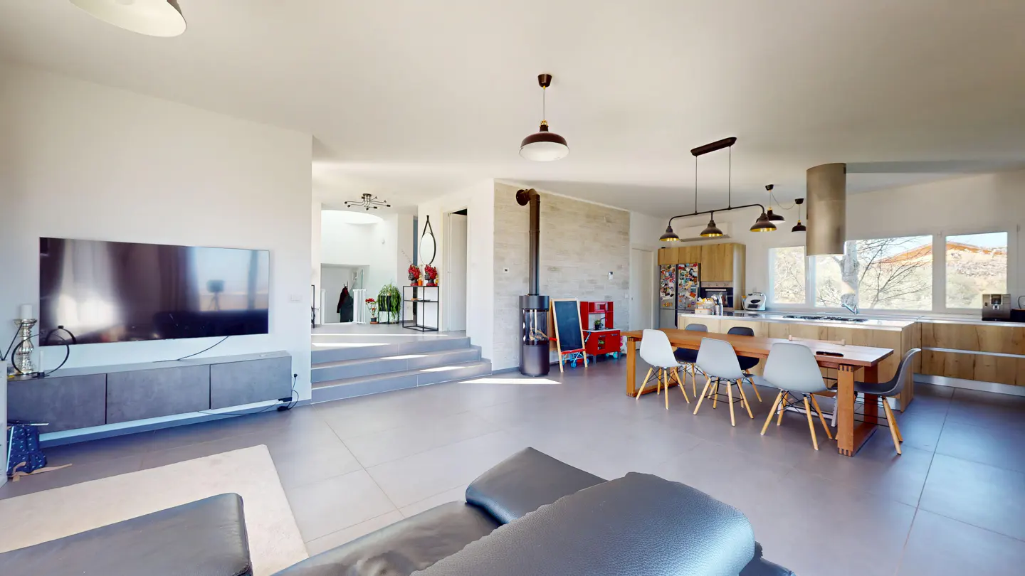 Open-concept living space with gray tile floors, a wood dining table, and a modern kitchen with light wood cabinets. A black wood stove stands near the dining area.