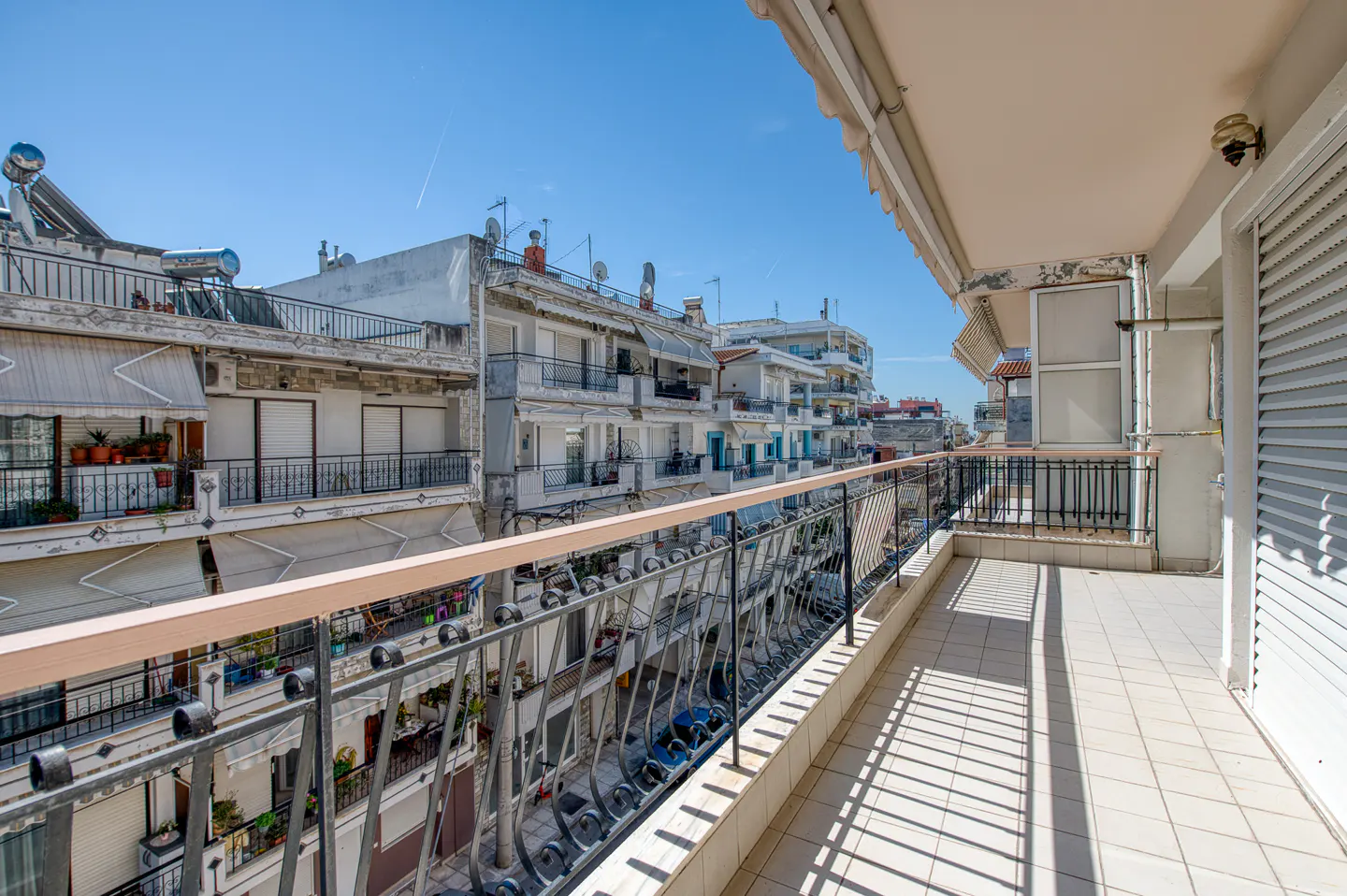View from a tiled balcony with black iron railings, overlooking a city street with buildings under a blue sky.