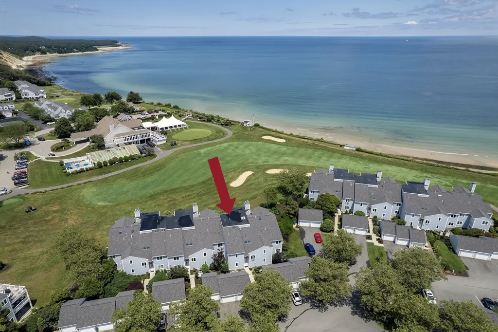 Aerial view of light blue townhouses with gray roofs, near a golf course and ocean, indicated by a red arrow.
