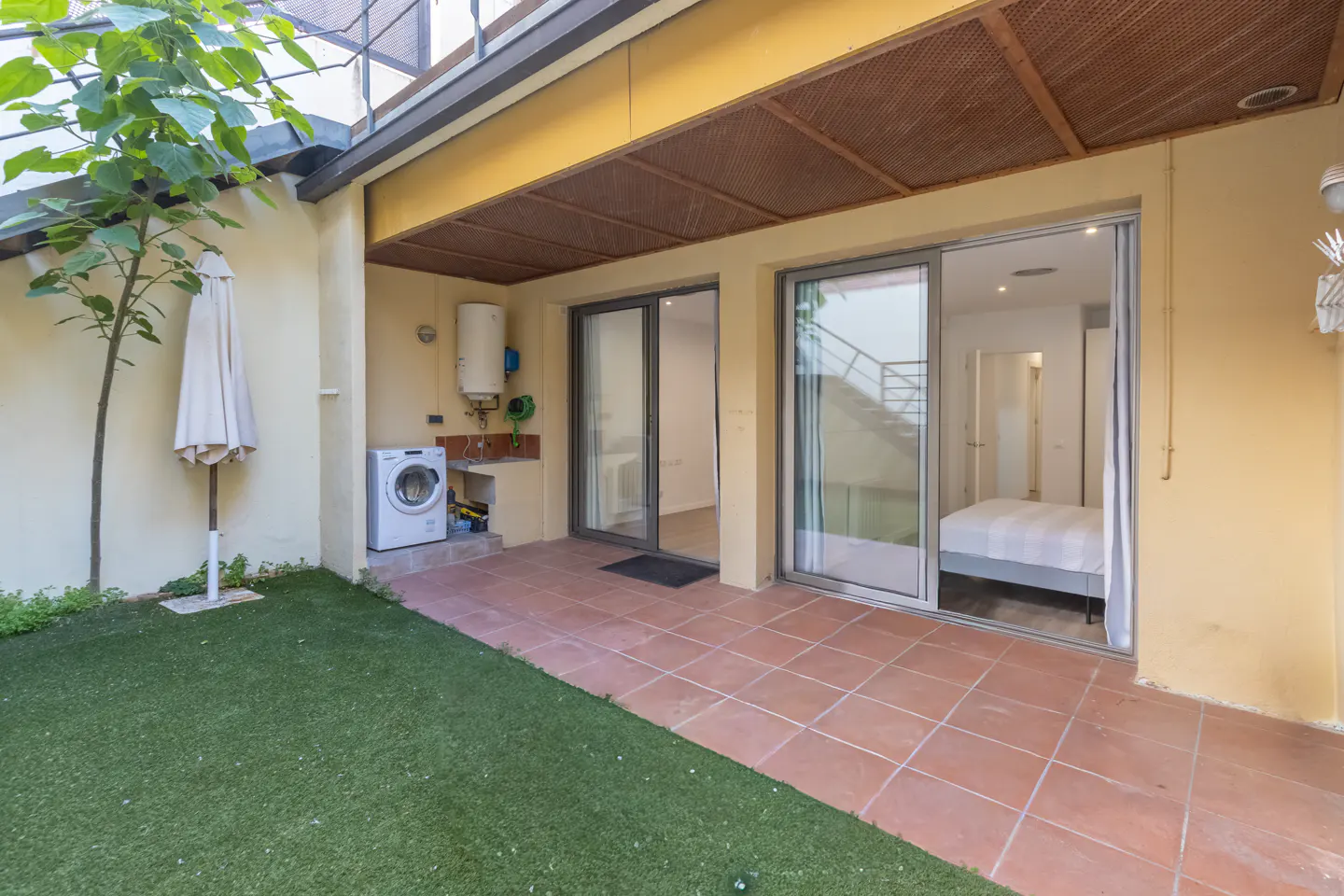Outdoor patio with terracotta tiles, green grass, washing machine, and sliding glass doors leading to a bedroom.
