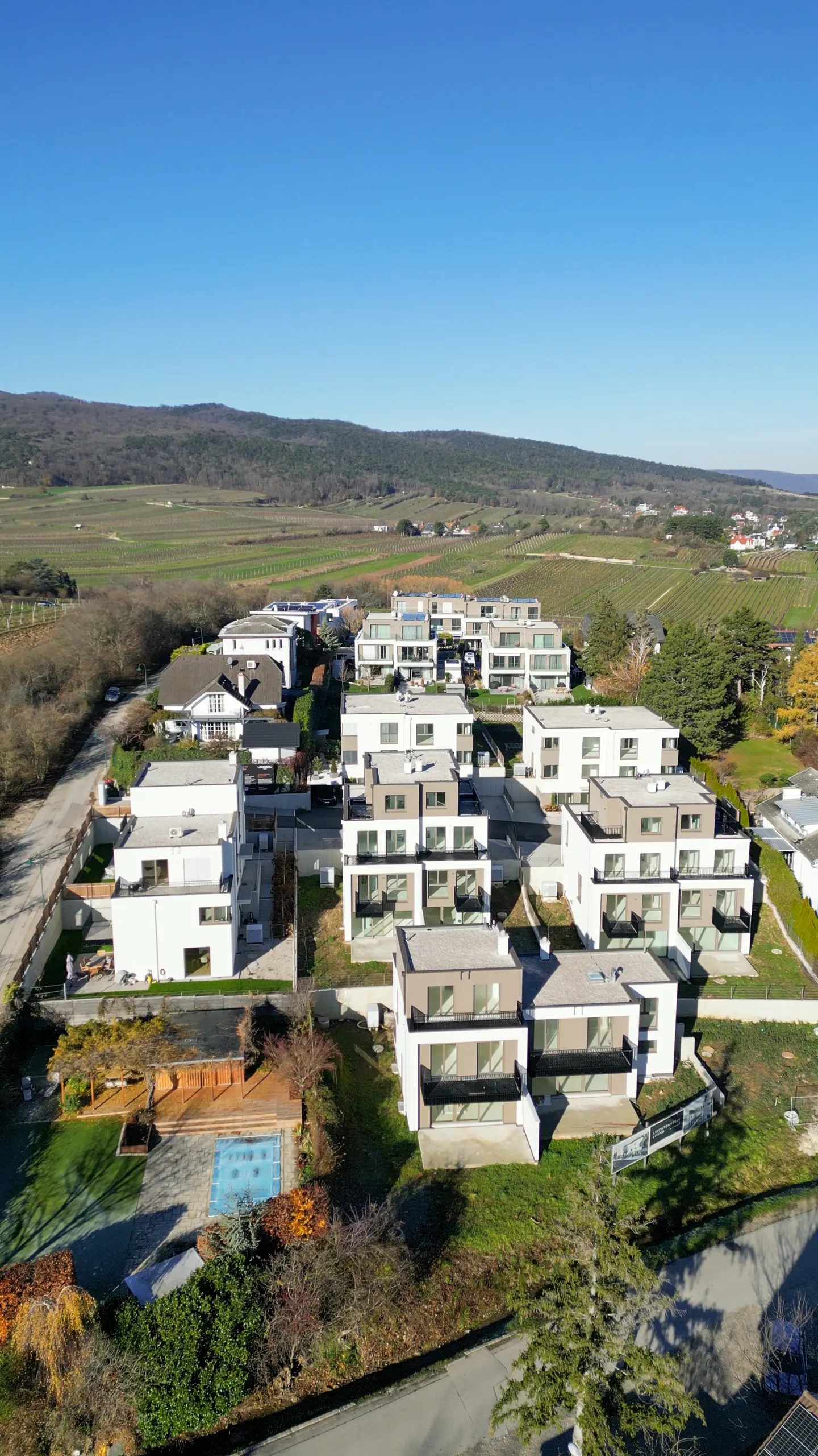 Aerial view of modern white houses with balconies, surrounded by green lawns and vineyards under a clear blue sky.