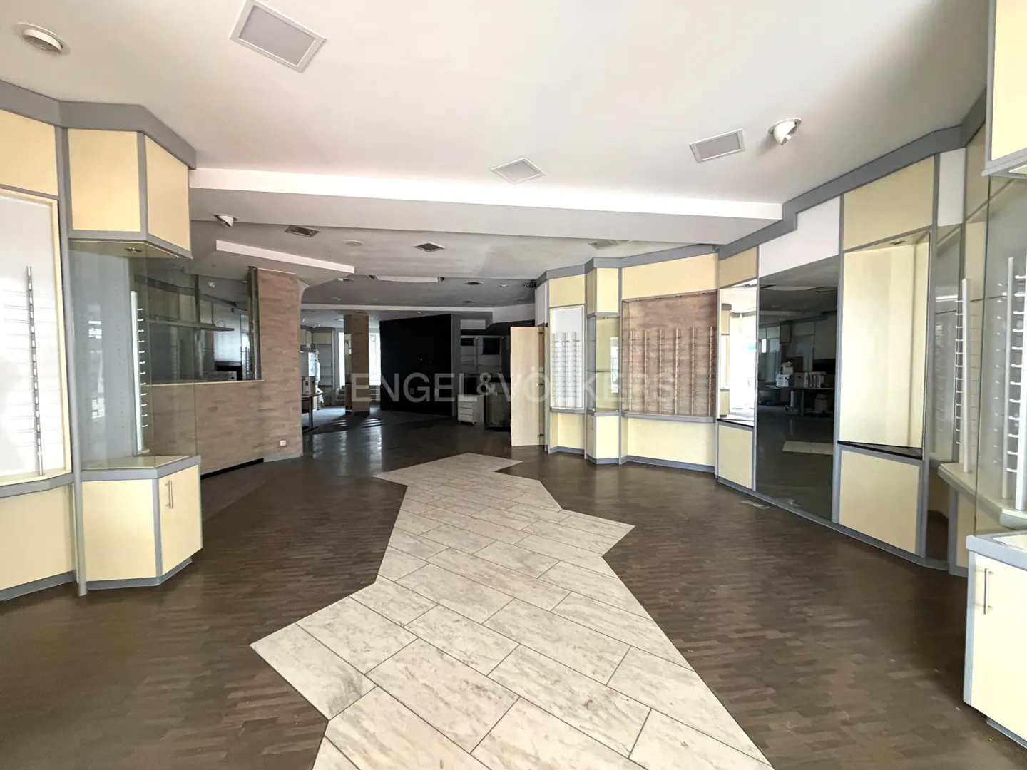 Empty retail space with beige walls, display cases, and a light tile path on a dark floor. Engel & Völkers logo visible in the background.