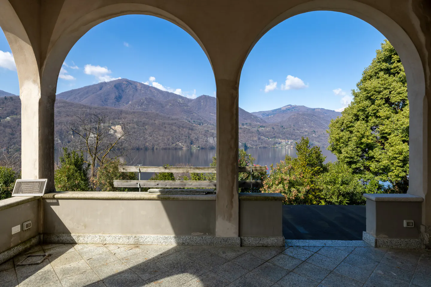 View from arched terrace with stone floor, overlooking lake, mountains, trees, and blue sky. A wooden bench sits on the terrace.