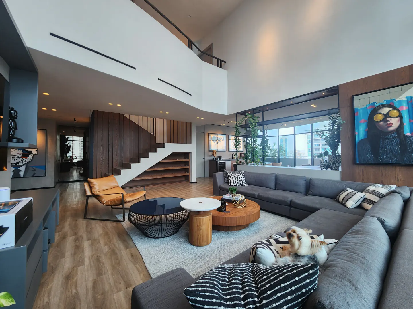 A modern living room with a gray sectional sofa, wooden floors, and a staircase. Artwork and a dog add personality to the space.