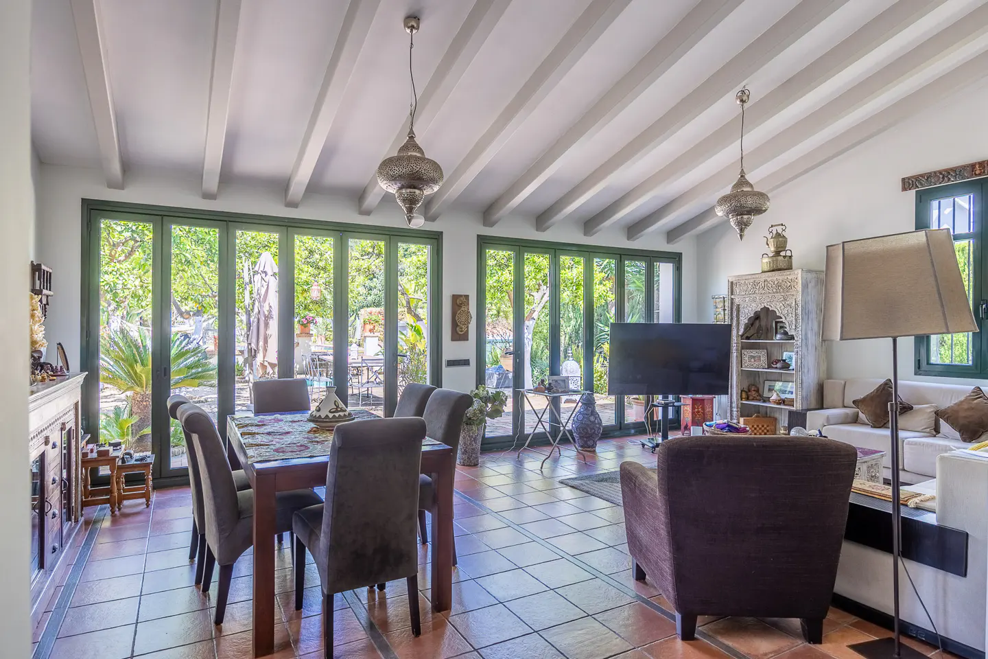 A bright living and dining area with terracotta tile floors, dark wood furniture, and folding glass doors to a lush garden.