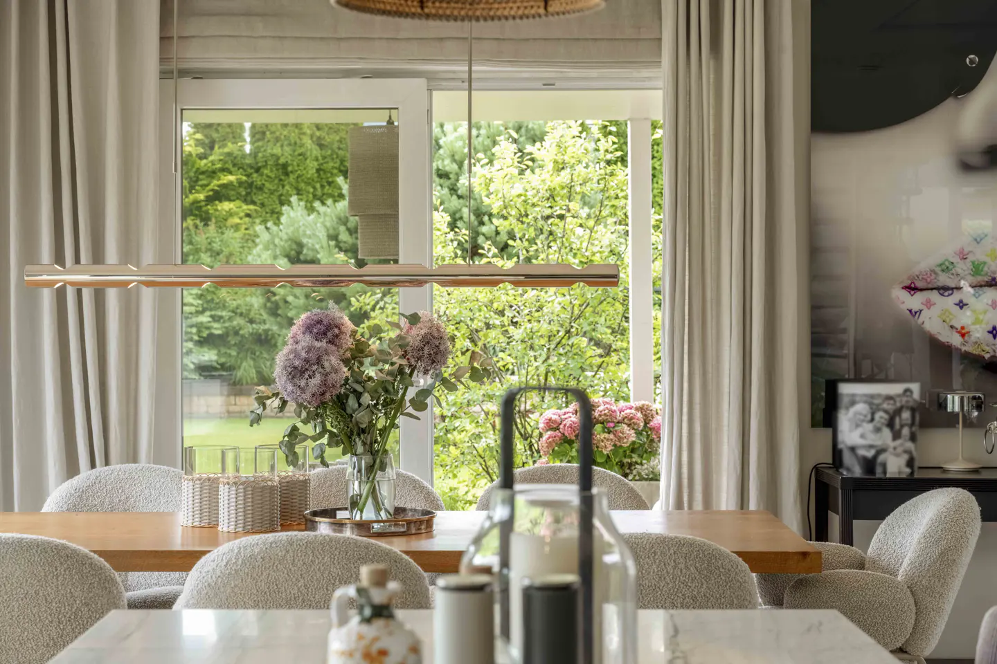Dining room with a wood table, beige chairs, and a vase of purple hydrangeas. A window overlooks a green garden.