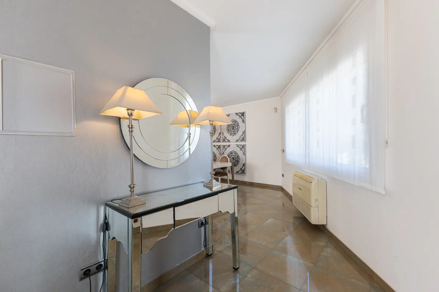 Entryway with a mirrored console table, lamps, and a round mirror on a gray wall. A hallway leads to a chair and patterned wall art.