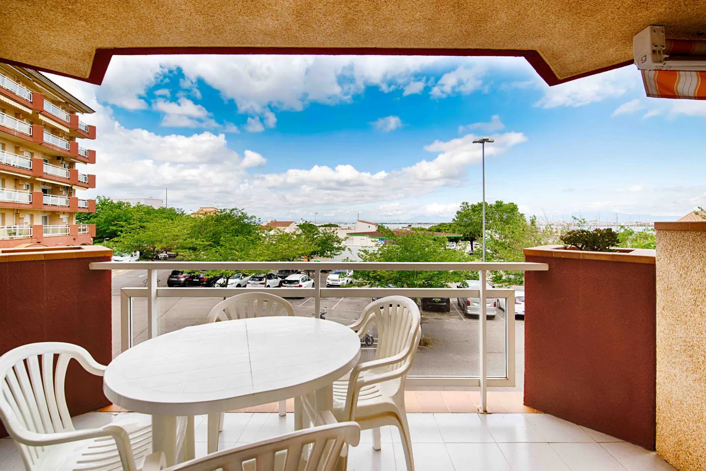 Balcony view with white table and chairs overlooking a parking lot, trees, and a blue sky with clouds.
