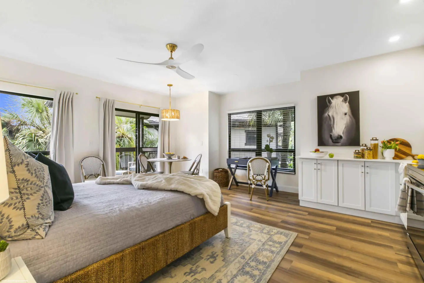 Bright studio apartment with a bed, dining table, and kitchenette. A horse portrait hangs above white cabinets. Windows show palm trees.