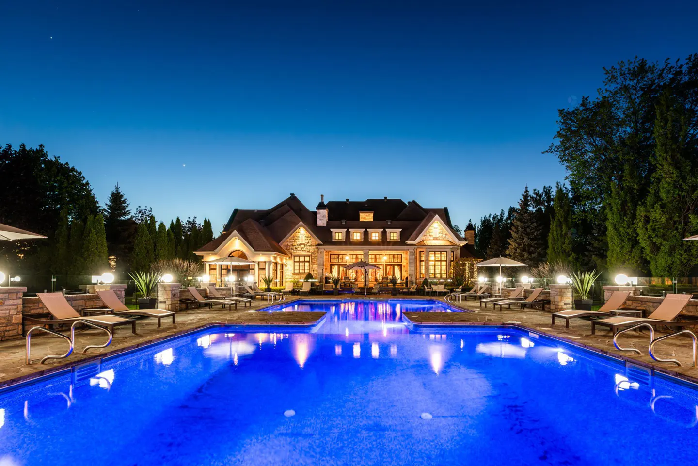 Night view of a large stone house with a blue-lit pool, lounge chairs, and trees.
