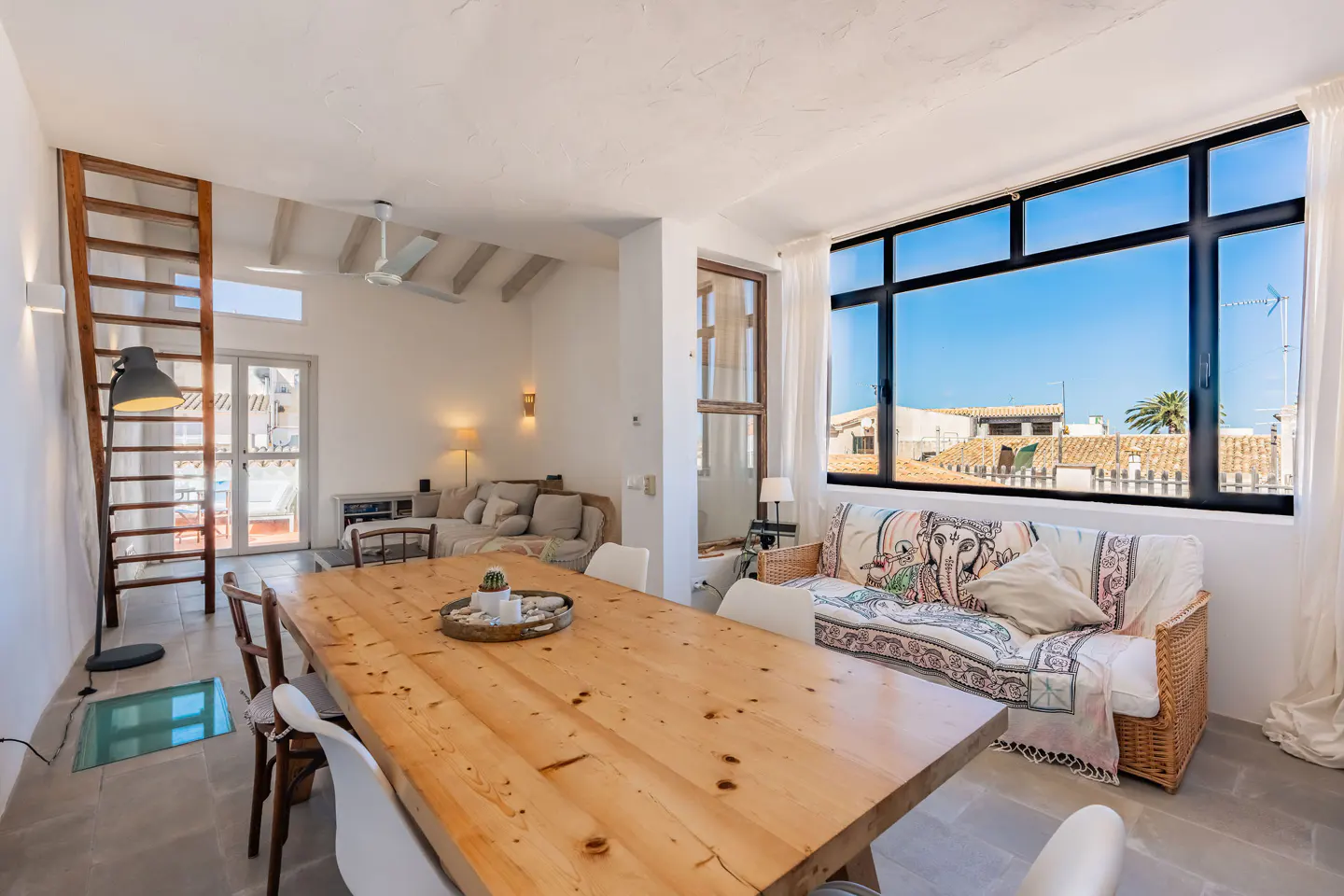 Bright, airy living space with a large wooden table, white chairs, and a wicker sofa by a window with a city view. A wooden ladder leads to an upper level.