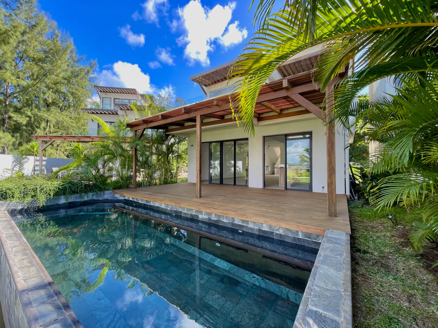 A modern white house with a wooden deck and a pool in the foreground, surrounded by lush greenery.