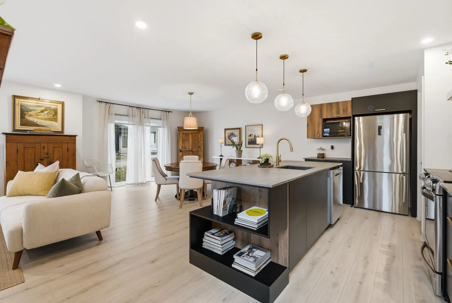 Open-concept living space with a kitchen island, dining table, and sofa. The kitchen features stainless steel appliances and modern lighting.