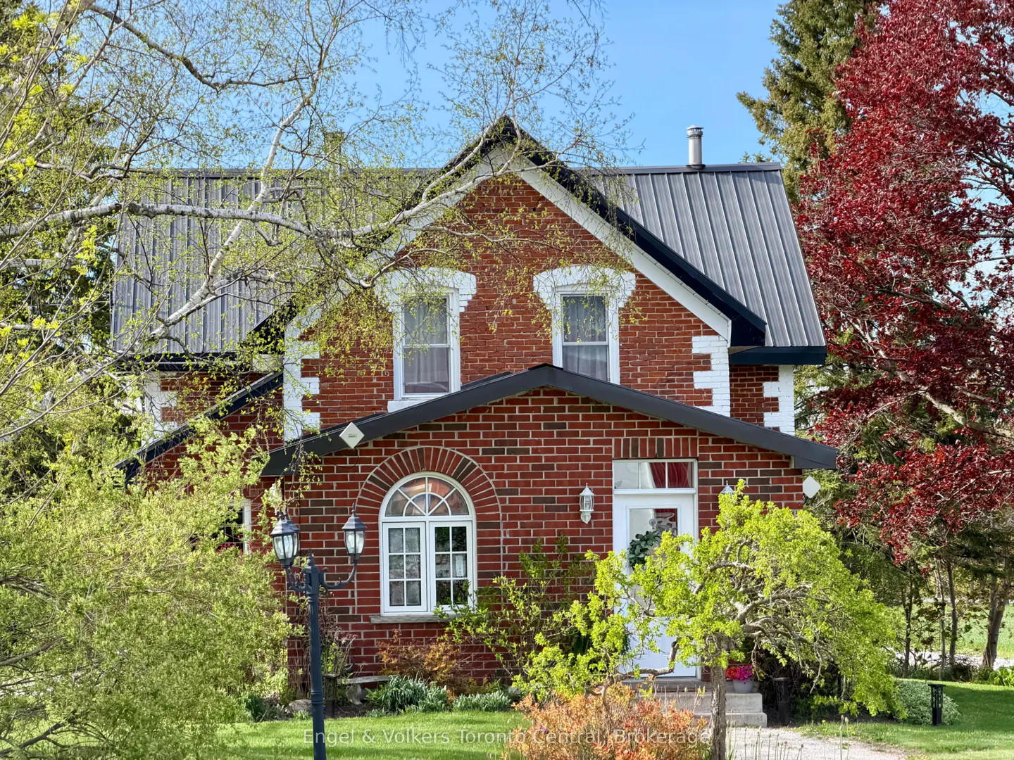 Two-story red brick house with a gray metal roof, white trim, and green trees.