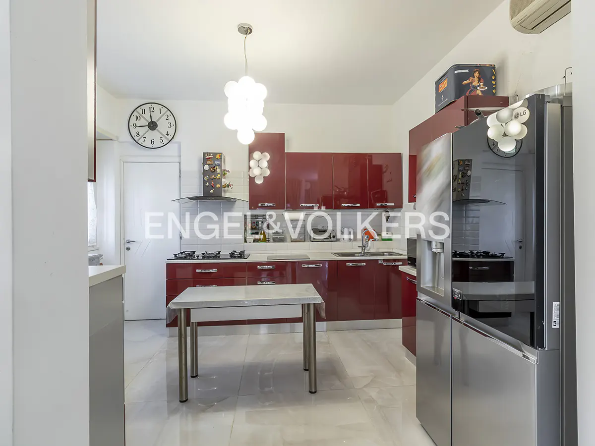 Bright kitchen with red cabinets, stainless steel fridge, and white marble floors. A white table stands in the center. A clock hangs on the wall.