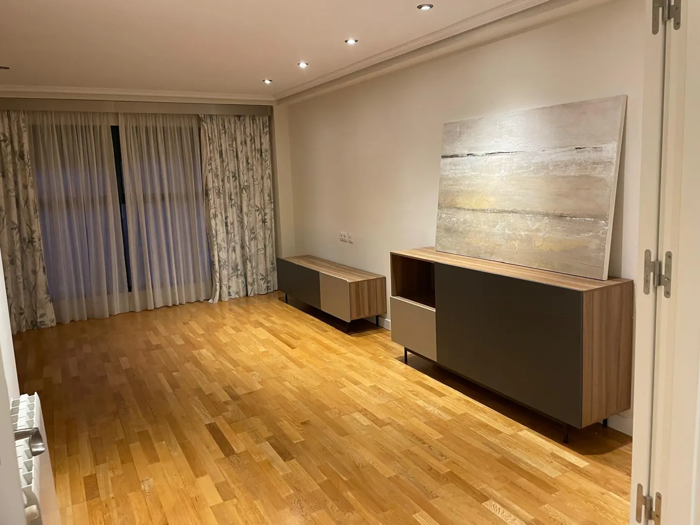 Living room with hardwood floors, beige walls, and recessed lighting. A large window with floral curtains is on the left. Two cabinets with an abstract painting are on the right.