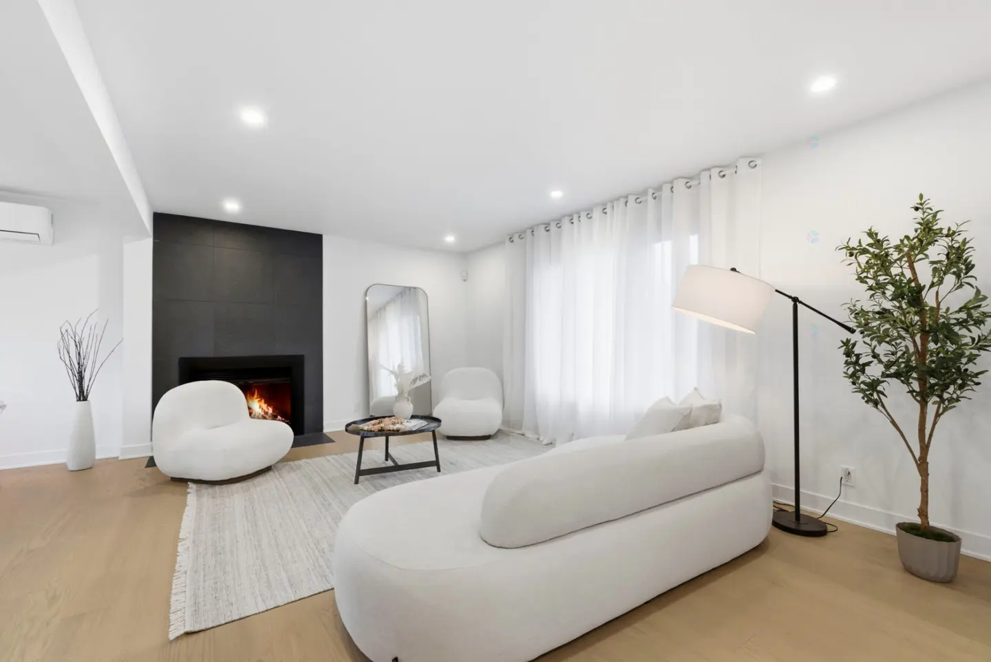 Bright living room with a black fireplace, white furniture, wood floors, and a large window with sheer white curtains.