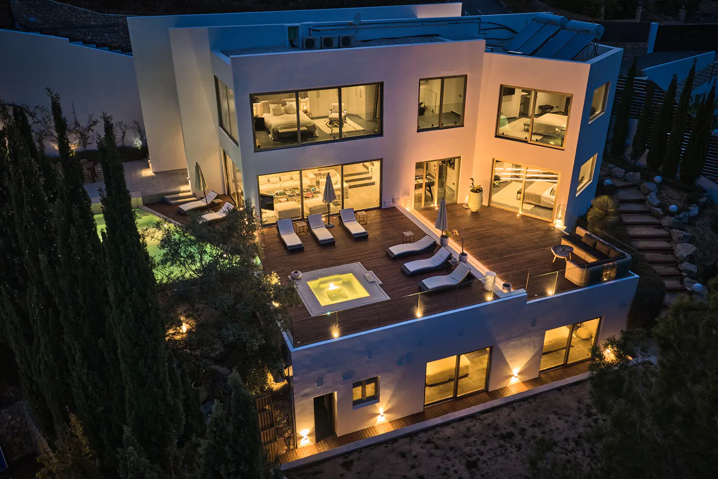 Aerial view of a modern, white, multi-story house at night. A wooden deck features lounge chairs and a lit hot tub. Large windows reveal interior bedrooms.