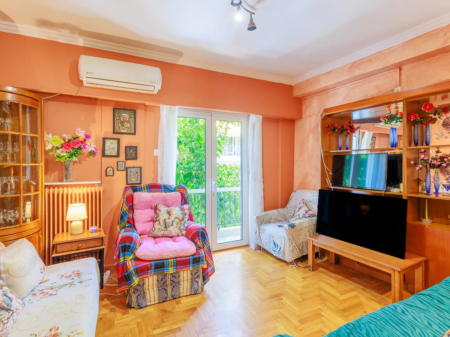 Living room with peach walls, hardwood floors, a plaid armchair, a TV on a wooden stand, and a balcony door with curtains.