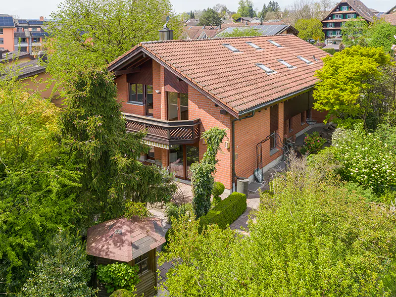 Aerial view of a two-story brick house with a red tile roof, surrounded by lush green trees and a small gazebo.