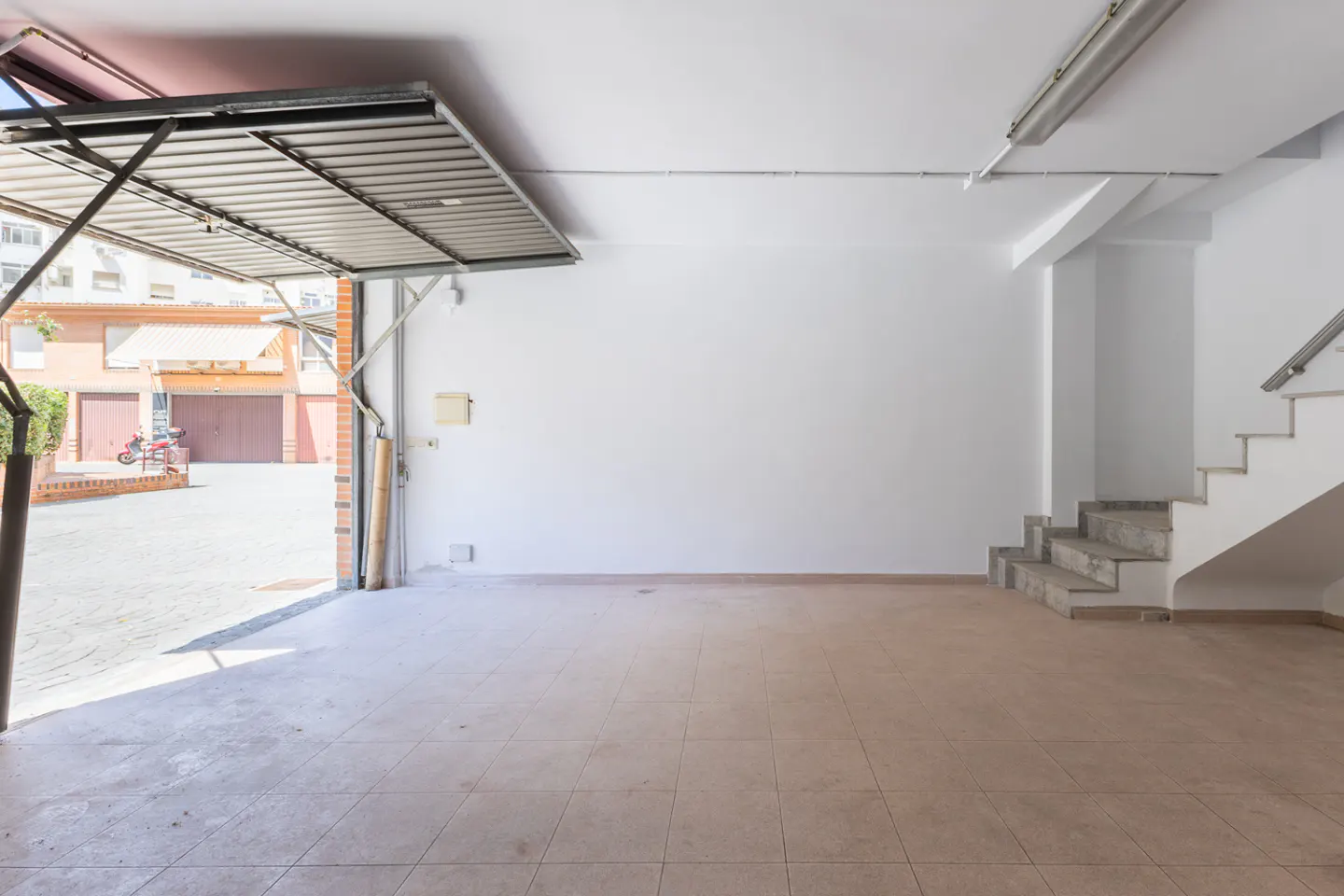 A bright, empty garage with an open metal door, beige tiled floor, white walls, and concrete stairs.