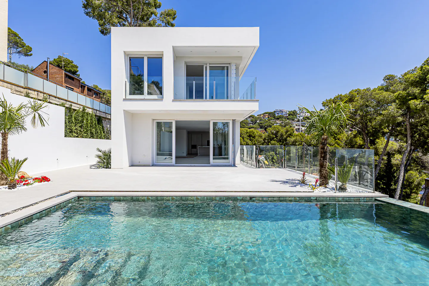 Modern two-story white house with a turquoise swimming pool in the foreground. Palm trees and green foliage surround the property.