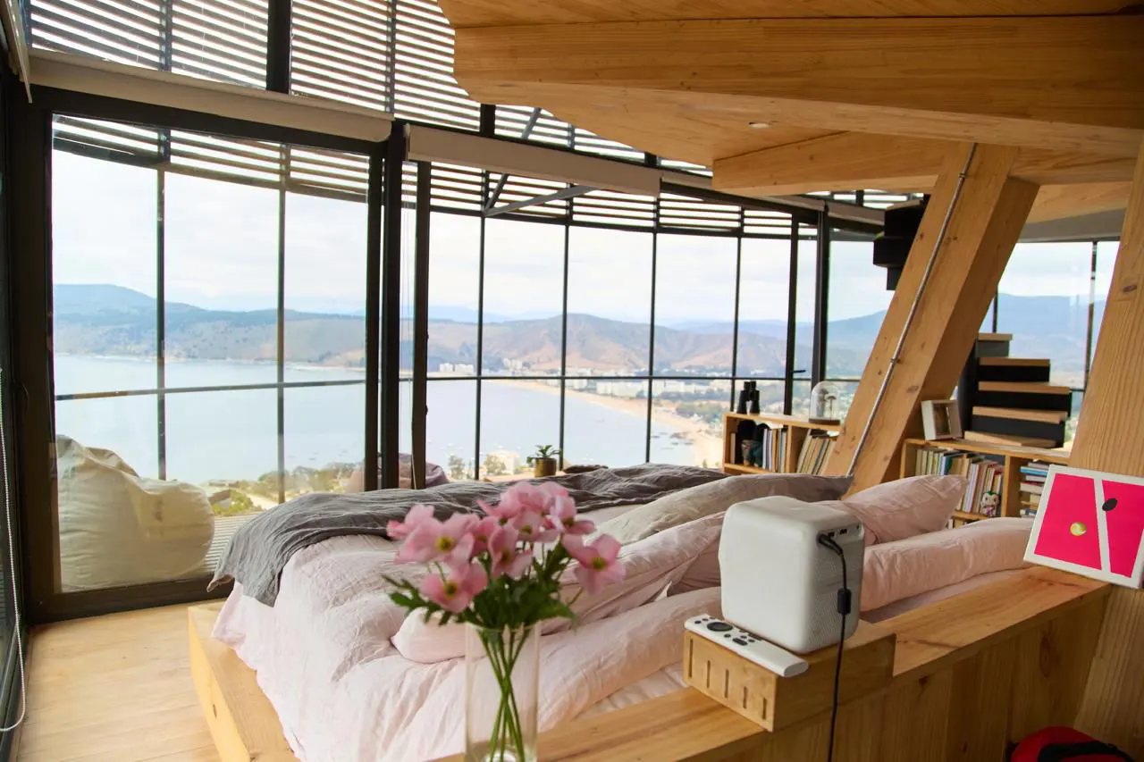 Bedroom with a large window view of the ocean. The room has a wooden frame, a bed with pink sheets, and a vase of pink flowers.
