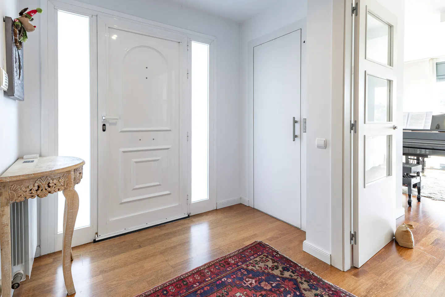 Bright entryway with white walls, a wooden floor, and a red and blue rug. A carved wooden table sits near the white front door.