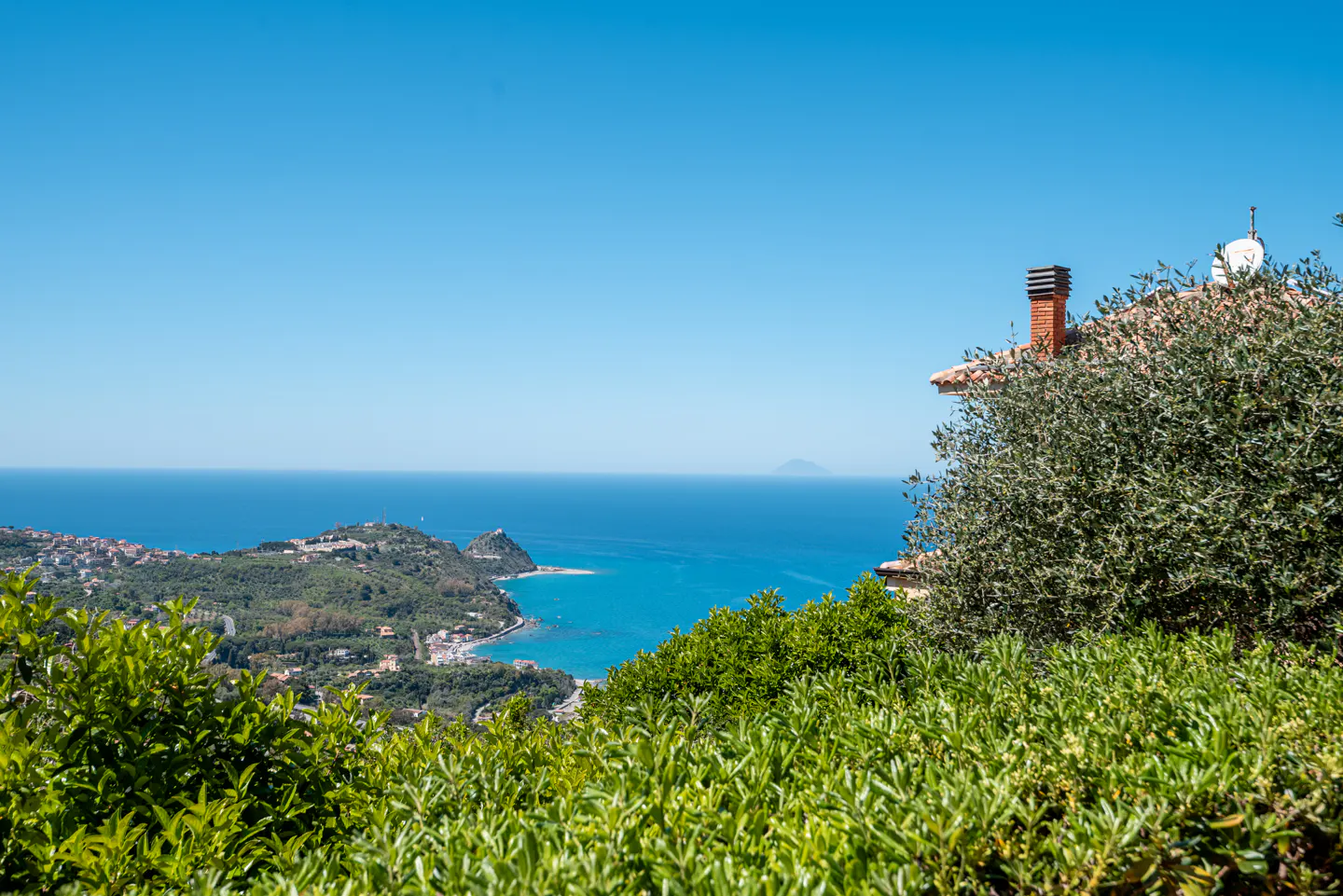 Scenic view of the Italian coastline with turquoise water, green hills, and a house with a red brick chimney on the right.