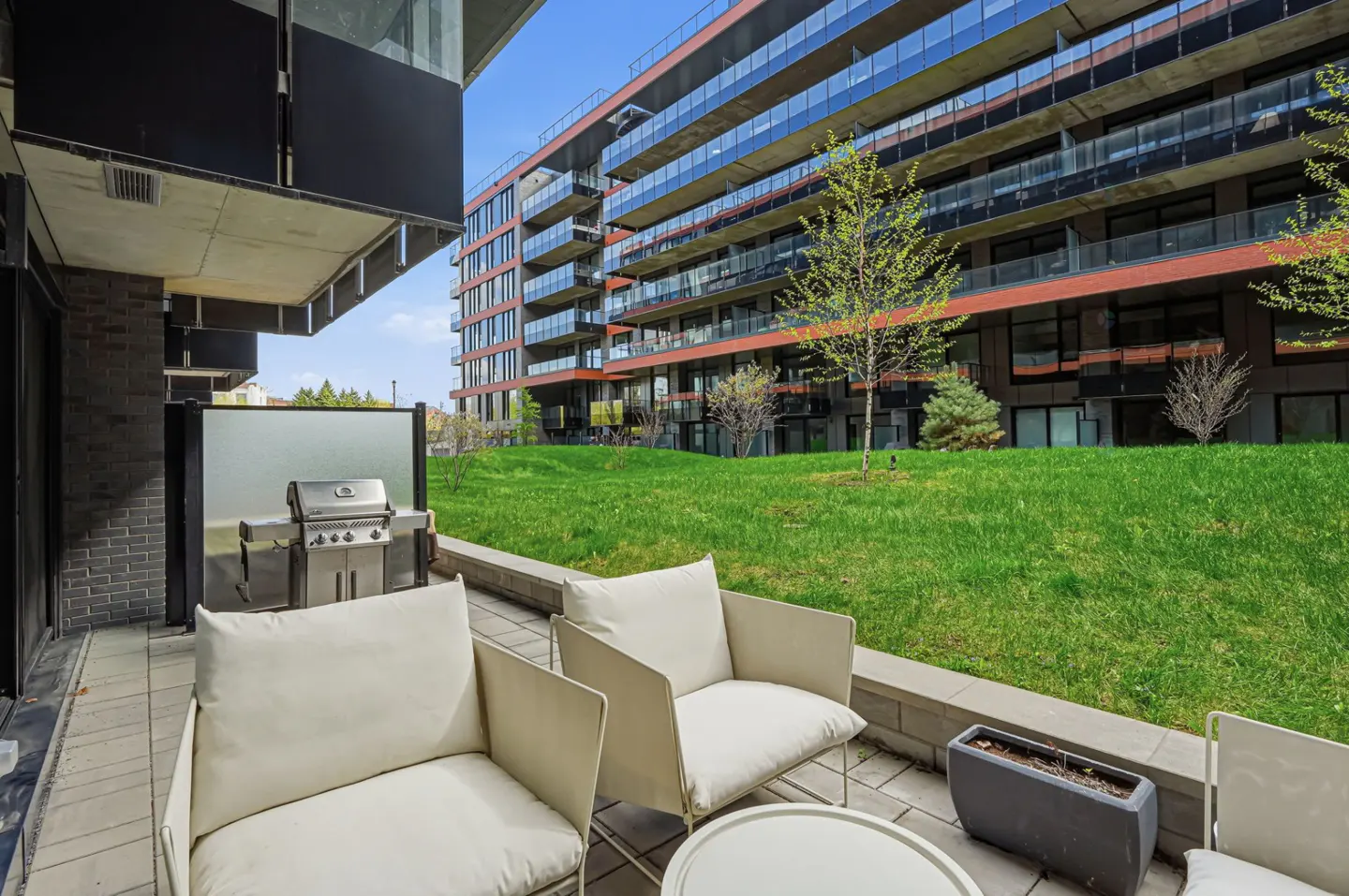 Condo patio with white chairs, table, and grill. A grassy lawn and modern building are in the background.
