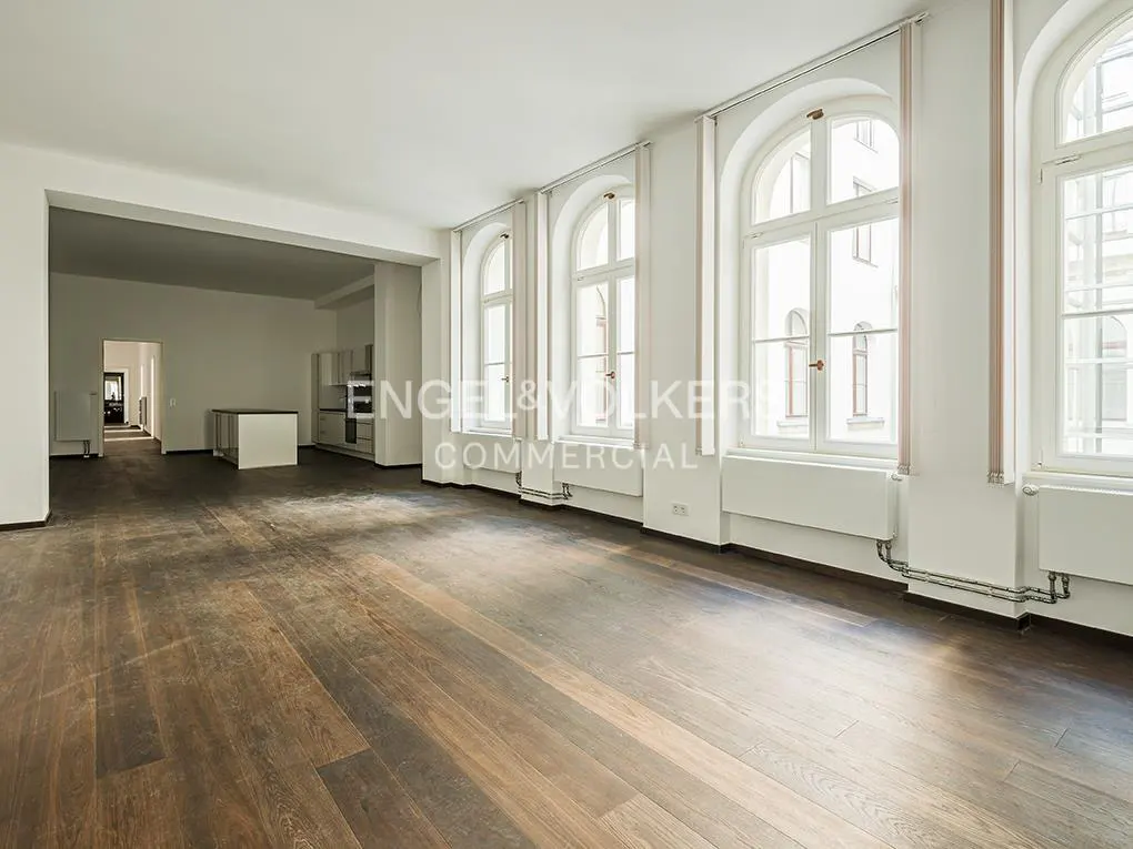 Bright, empty room with dark wood floors, white walls, and arched windows. A kitchen area is visible in the background.