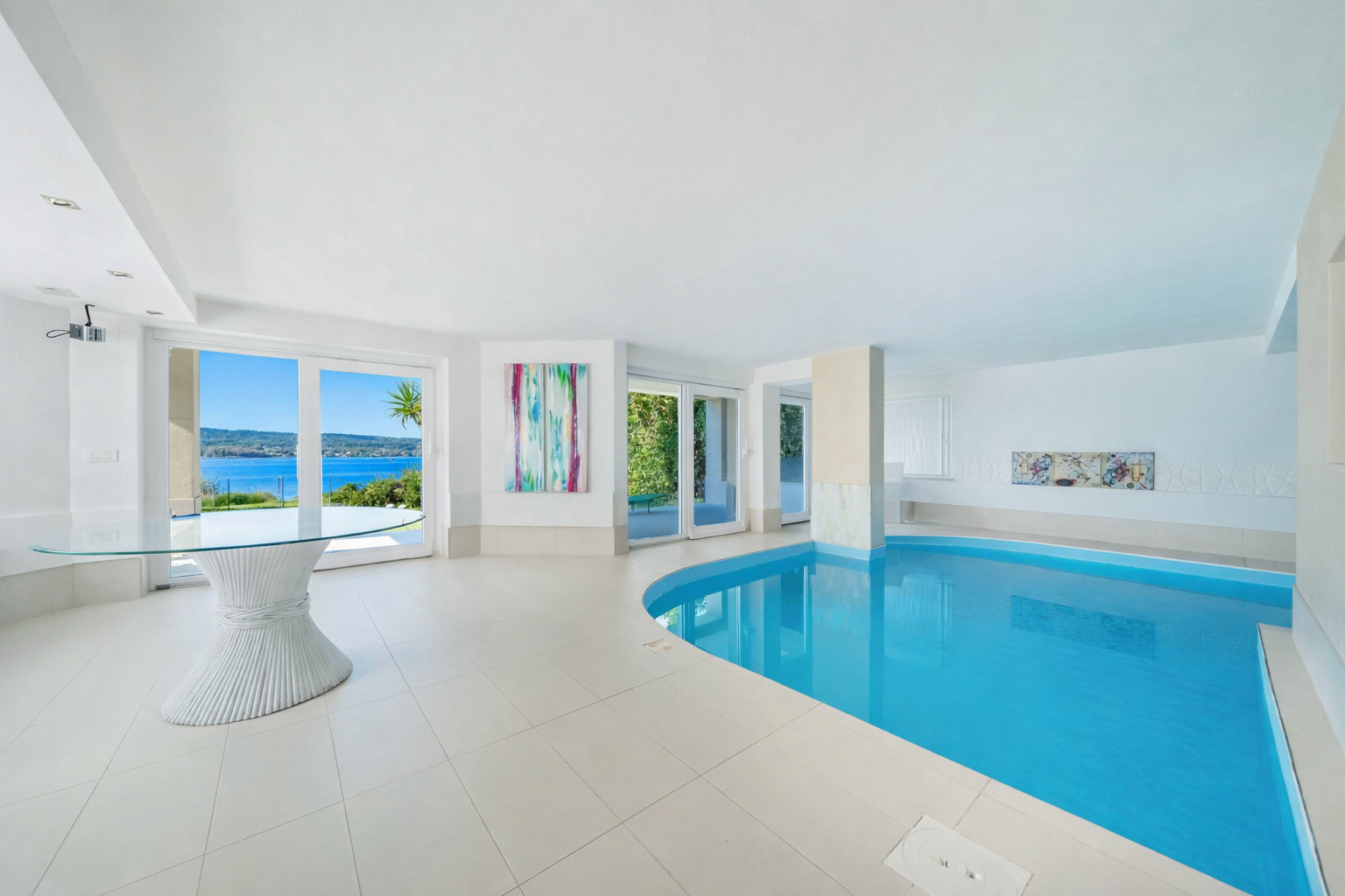 Bright indoor pool room with white tile floors, a glass table, and large windows overlooking a blue lake and sky. [AI-generated text]