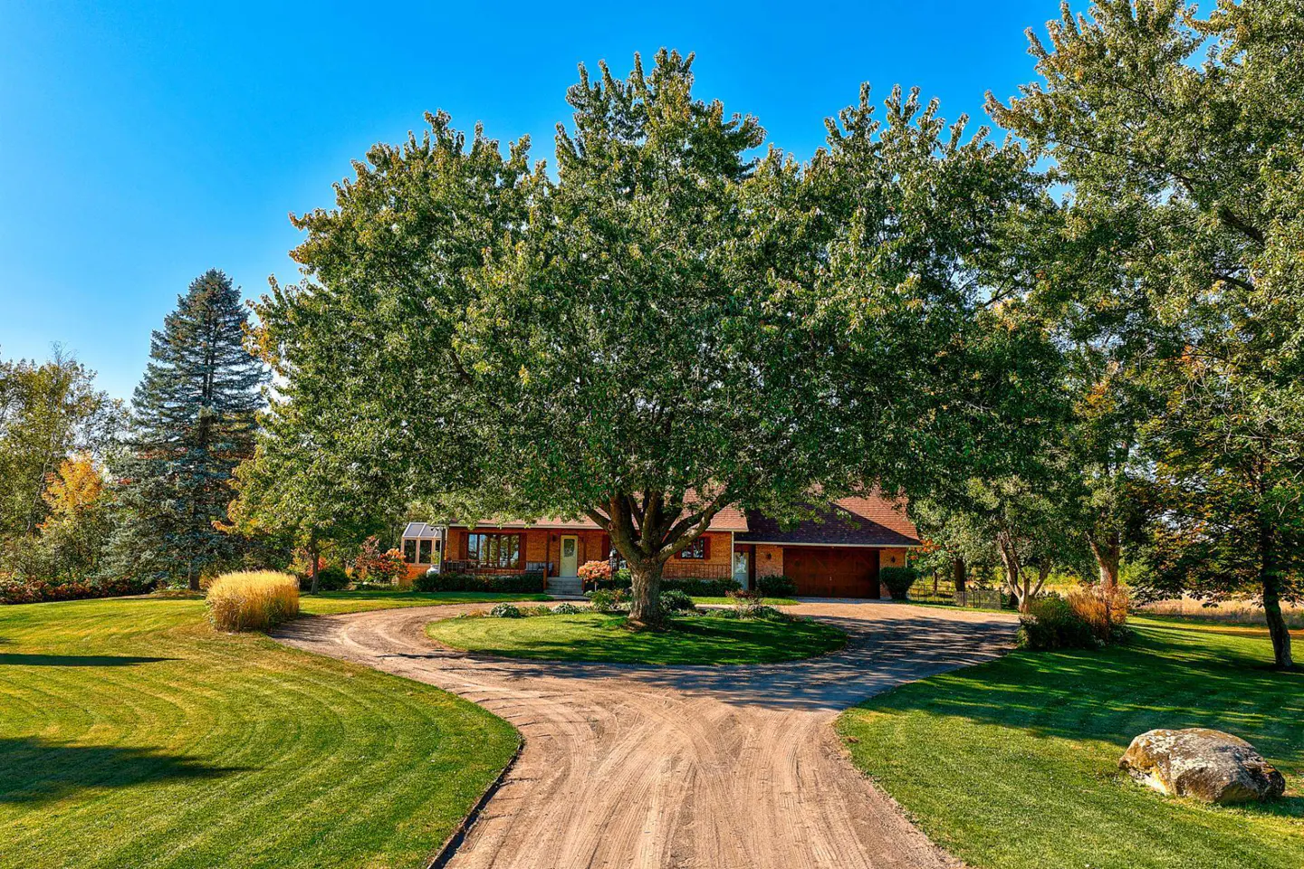 A long driveway leads to a brown house, partially obscured by a large green tree, under a clear blue sky.
