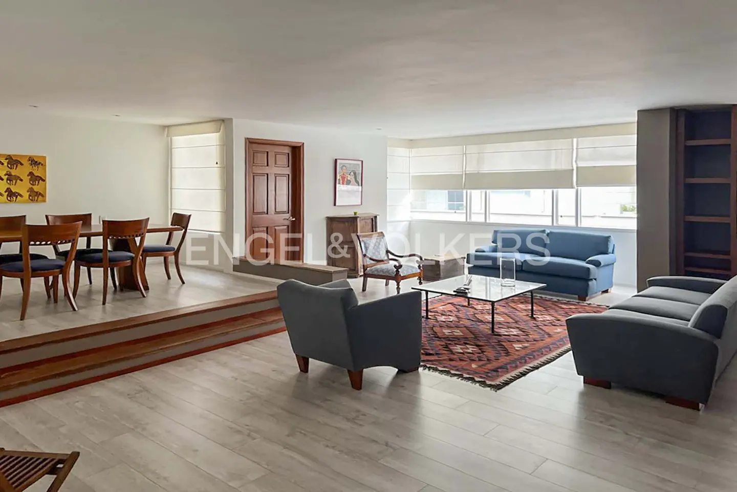Spacious living and dining area with light wood floors, blue sofas, a patterned rug, and a raised dining room section.