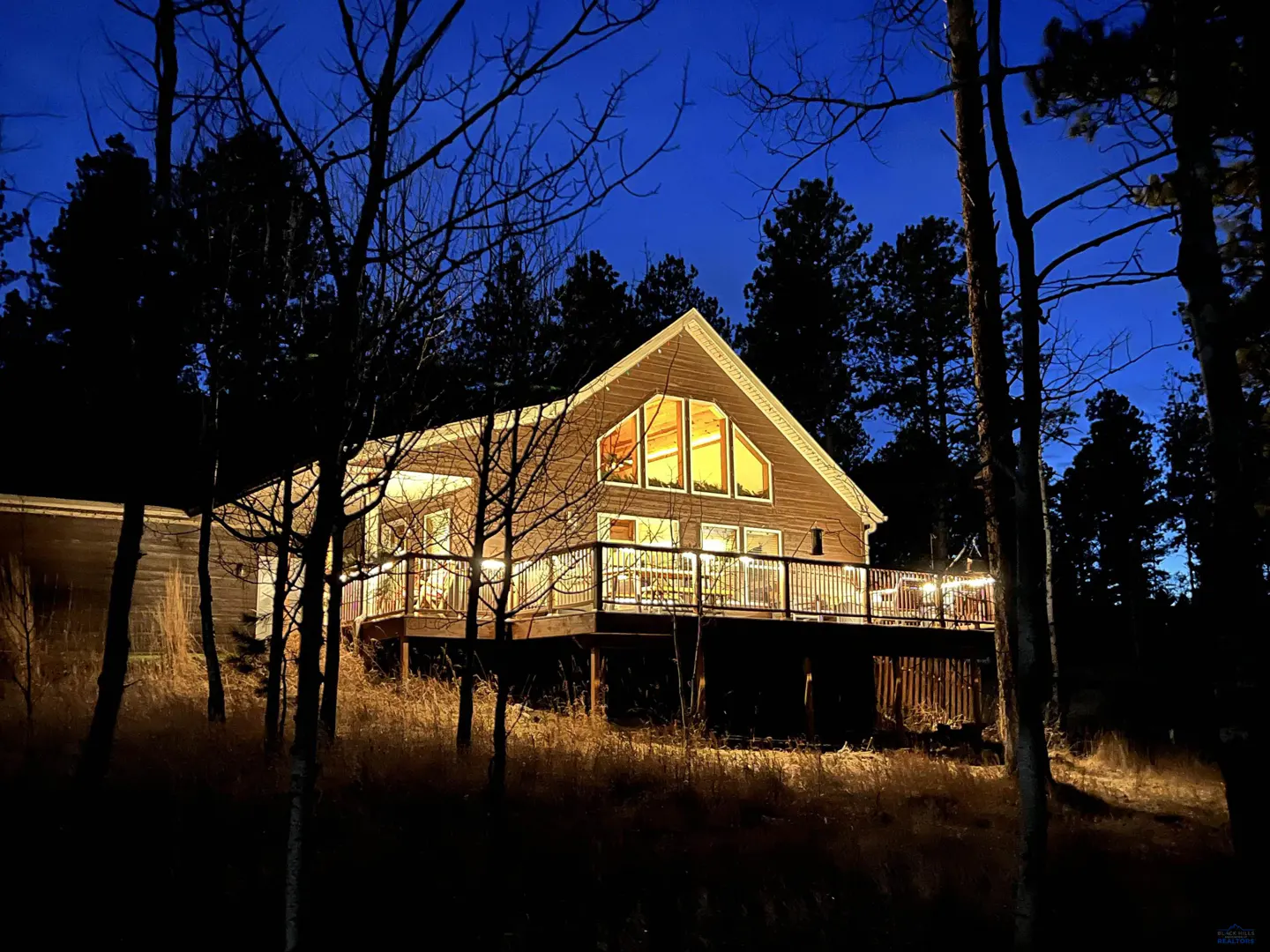 A wooden house with a lit deck and large windows glows against a dark blue twilight sky, surrounded by silhouetted trees.