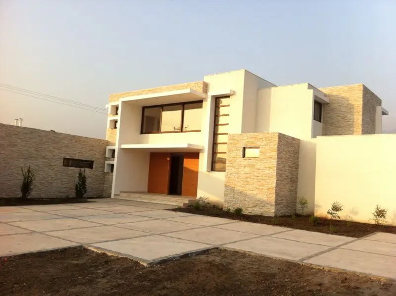 Modern two-story house with white walls, stone accents, and a concrete driveway. A wooden front door is visible.