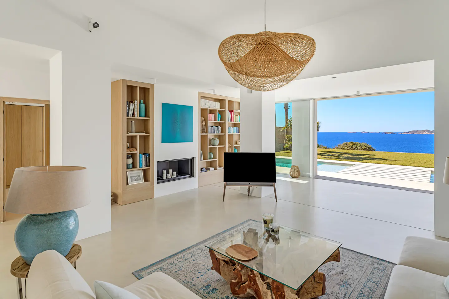 Bright, modern living room with white walls, wood shelves, and a glass coffee table. Ocean view through sliding glass doors.