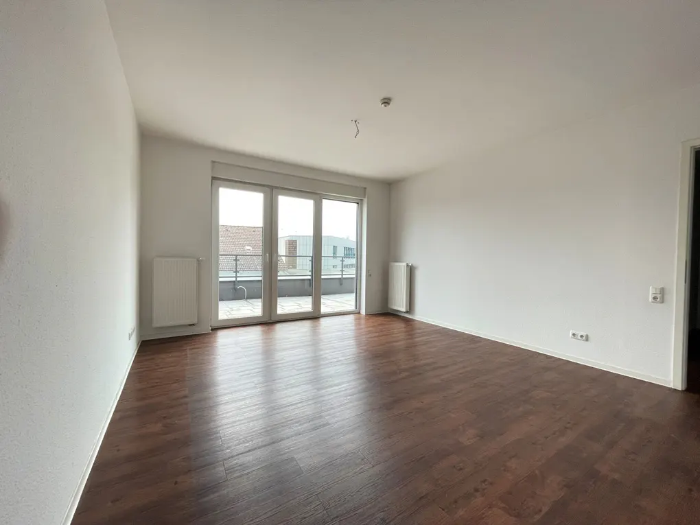 Bright, empty room with dark wood floors and white walls. Glass doors lead to a balcony. Radiators are on the walls.