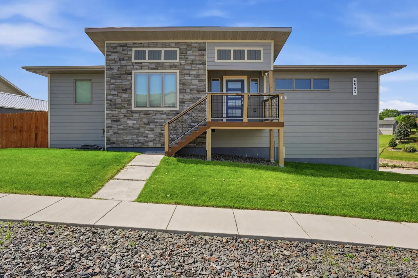 Modern gray house with stone facade, elevated front porch, and blue door under a bright blue sky.