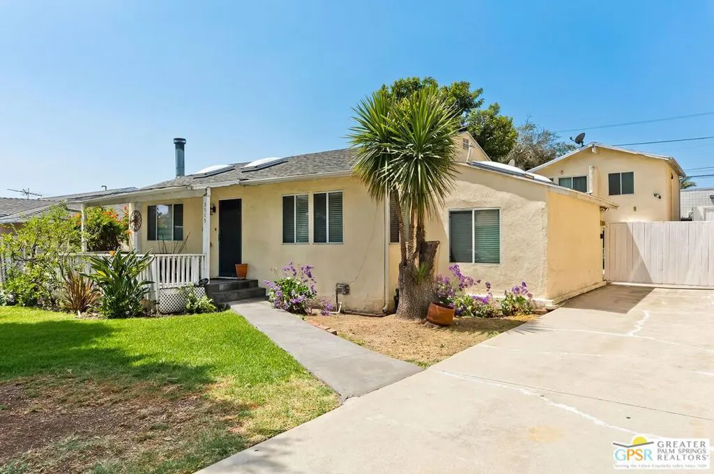 A one-story, light yellow house with a gray roof, a small porch, and a palm tree in the front yard.