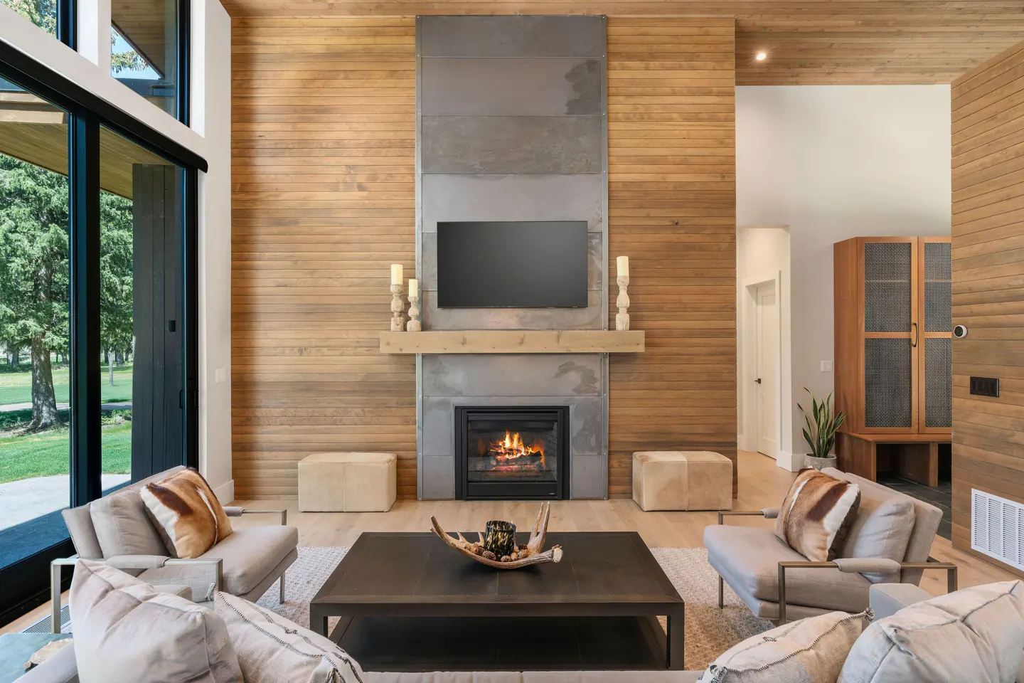 Living room with wood-paneled walls, a gray fireplace with a TV, and tan chairs around a dark coffee table. A large window shows trees.