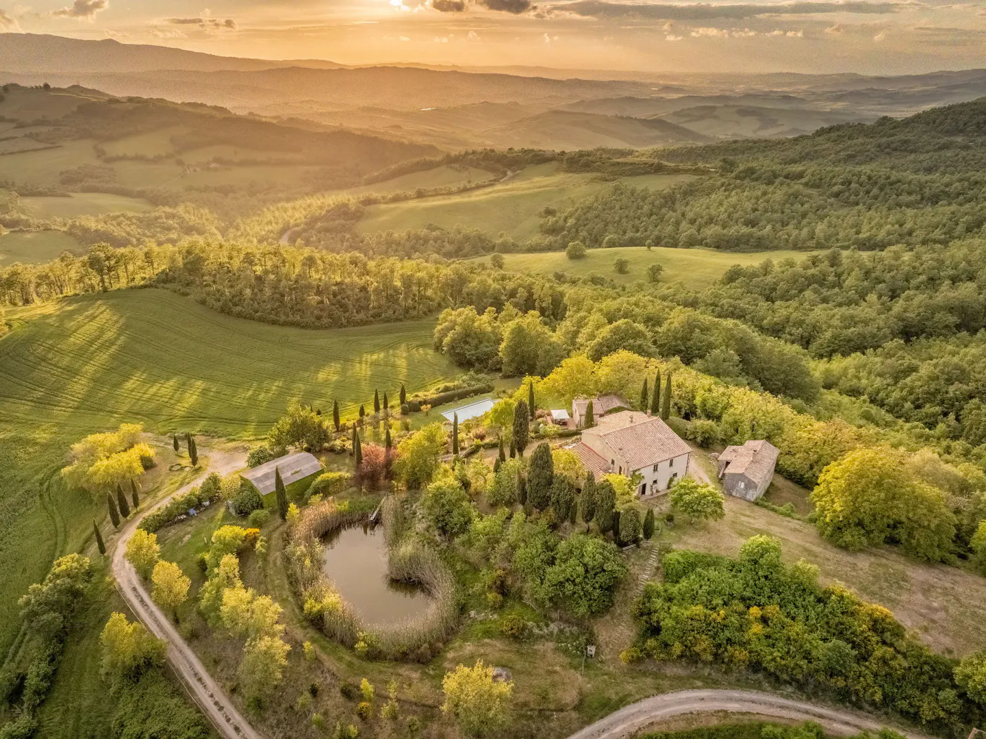 Aerial view of a Tuscan villa with a pond, pool, and rolling green hills at sunset.