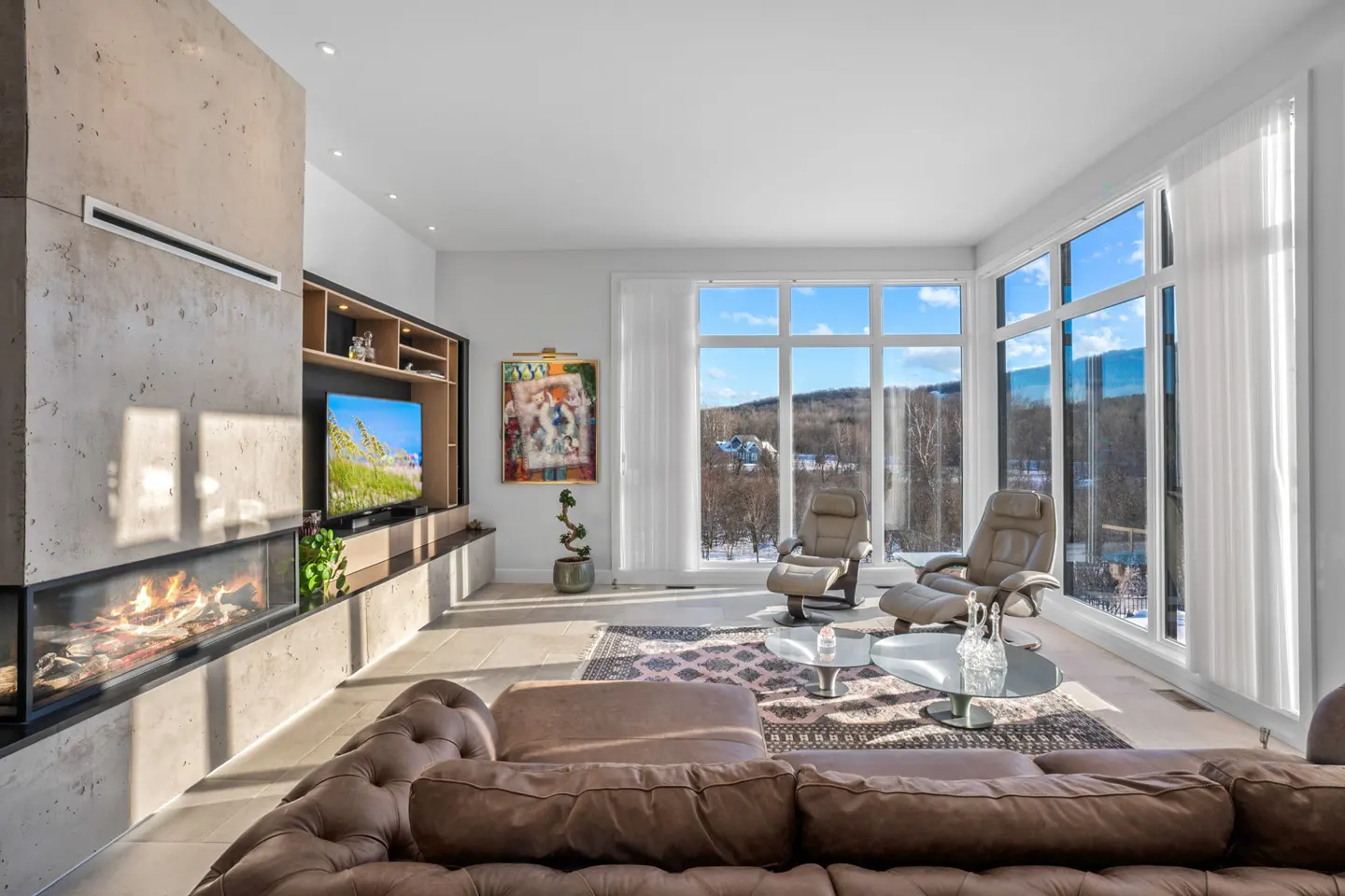 Bright living room with a brown leather sofa, fireplace, TV, and two recliners facing a large window with a winter view.